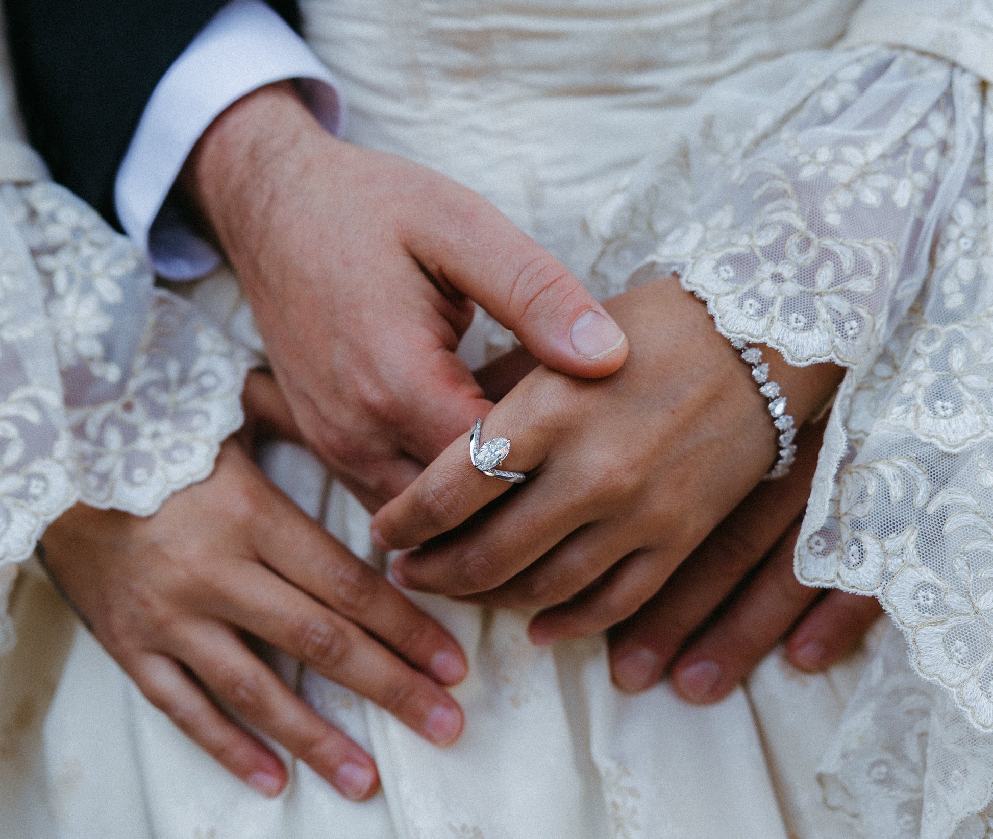 Close-up of hands holding each other with a wedding ring, wearing lace sleeves and a wedding dress.
